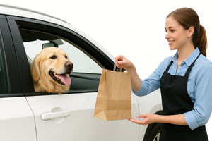 Une femme souriante en tablier tend un sac en papier à un golden retriever assis à l'avant d'une voiture blanche, fenêtre ouverte. Le chien a l'air heureux et tire la langue.